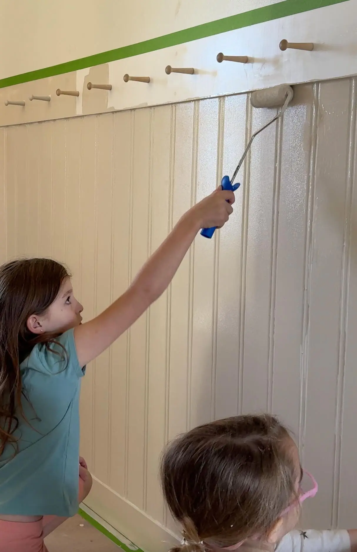 A young girl using a paint roller to cover beadboard paneling with paint, reaching up to paint a decorative wooden peg rail. Painter’s tape neatly lines the top of the wall, highlighting the effort to create clean edges in this DIY project.